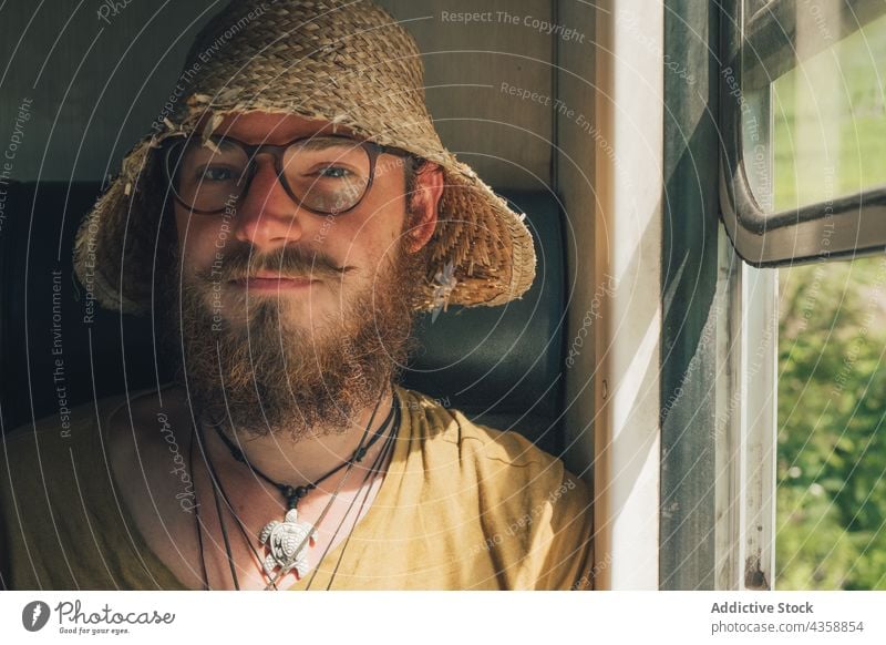 Content traveling man riding in train and looking at camera window hipster straw hat traveler smile explorer railway male railroad carriage wagon trip journey