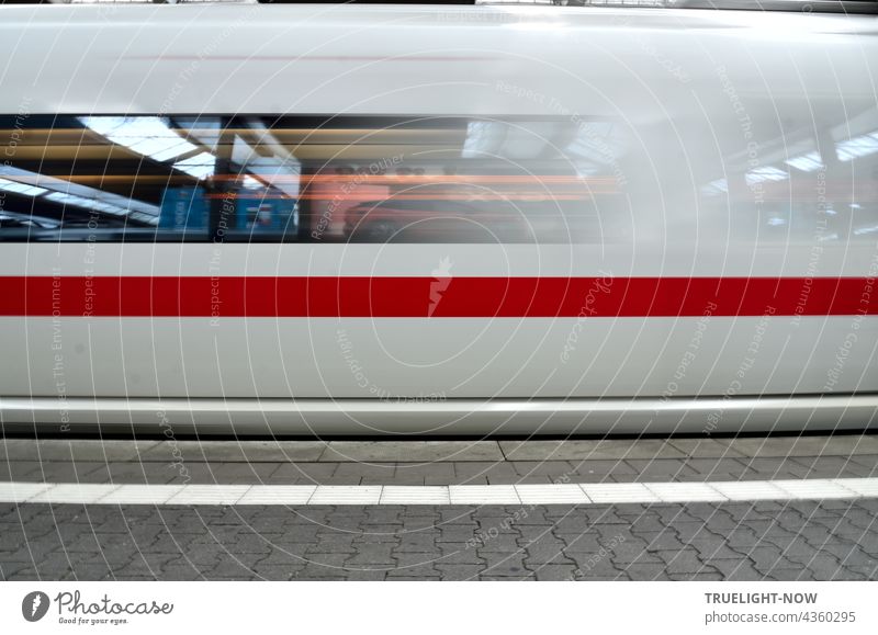 A snow-white ICE train with a thick red stripe is just leaving Munich Central Station. In its windows the station concourse is reflected with neon lights, the grey paving of the platform shows a white stripe