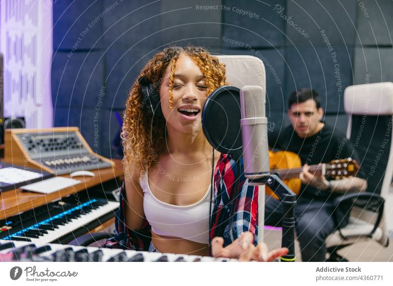 Cheerful multiethnic musicians playing guitar and synthesizer while singing indoors cheerful studio recording using gadget man woman device headphones acoustic