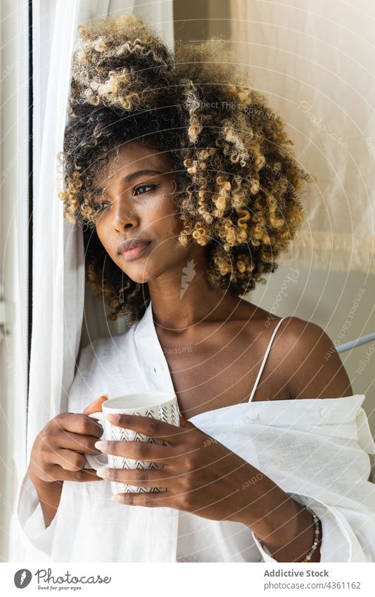 Dreamy black woman with cup of drink near window peaceful dreamy tranquil morning serene at home charming attractive female beverage brunette mug hot drink calm