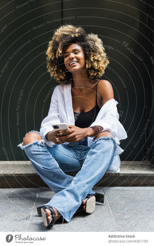 Smiling black woman using smartphone while sitting on concrete border positive internet enjoy surfing digital happy pleasant female cellphone legs crossed