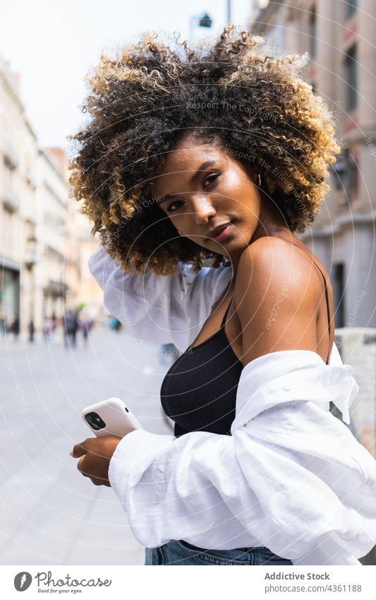 Black woman touching hair while standing on street with smartphone individuality cool serious urban millennial walkway posture female beautiful building device