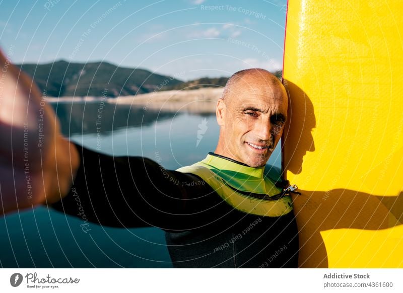 Smiling mature man with paddle board taking selfie on lake shore paddleboard activity practice smile sporty yellow male middle age water sports hobby active