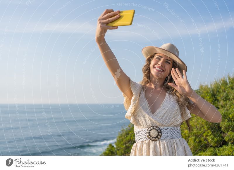 Happy woman taking selfie at seaside fashion summer style happy smartphone beach look vacation traveler female young cheerful holiday hat mobile smile wave hand