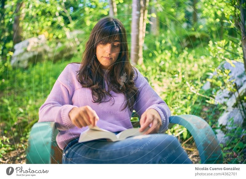 Relaxed latina woman reading a book sitting on a vintage turquoise chair, outside on her porch, surrounded of nature, trees and grass. relaxed young adult