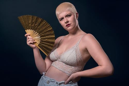 studio shot of a young, strong woman with very short blonde hair, a fan and a bustier serious confident power powerful business piercing jewelry portrait female