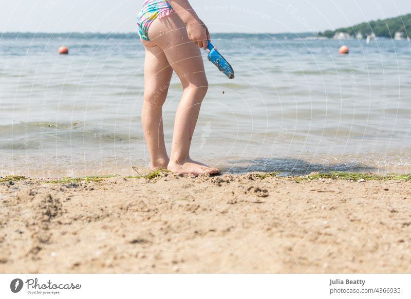 Little girl standing at the water's edge holding a blue plastic shovel; playing at the lake in the summer child swimsuit scoop build sand seaweed Midwest shadow
