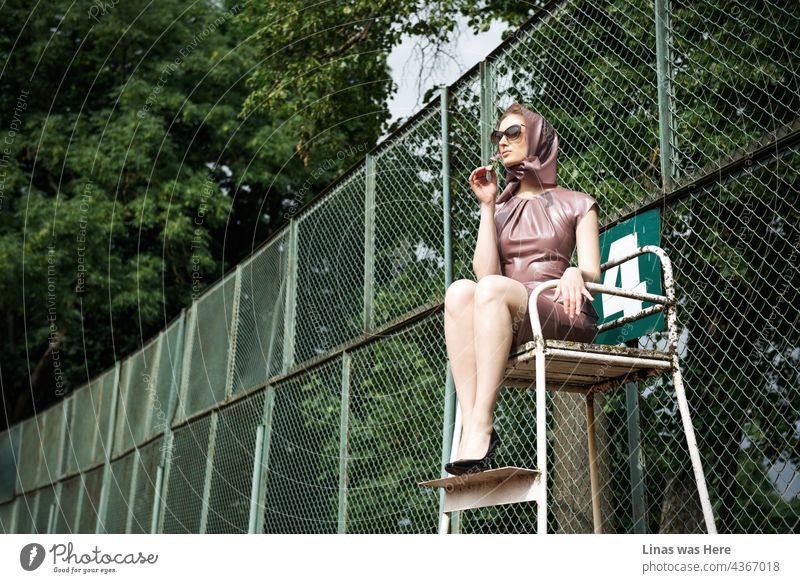 Next to a rusty fence of a tennis playground a gorgeous model is sitting all beautifully. She is wearing a pink latex dress, fashionable sunglasses, and high heels of course. My kind of judge doing all the pretty work.