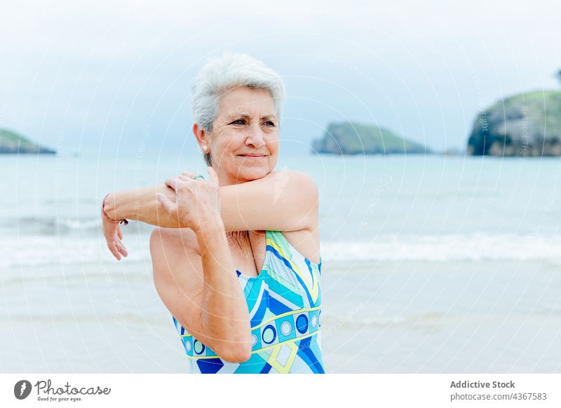 Happy senior woman exercising near ocean exercise workout beach healthy lifestyle stretch shoulder sea swimsuit arm activity wellbeing female gray hair old