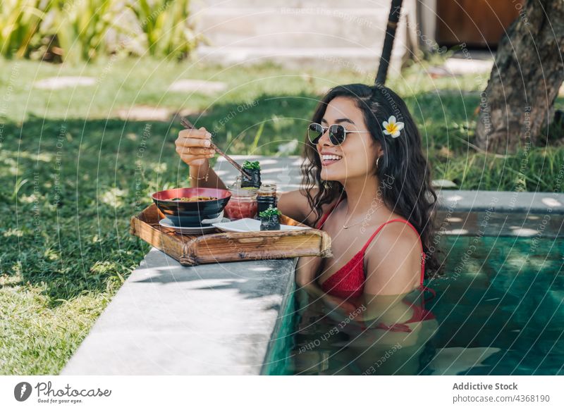 Smiling tourist with tasty noodles in pool during trip asian food sushi resort smile delicious woman chopstick swimwear traveler gunkan lunch seafood chuka