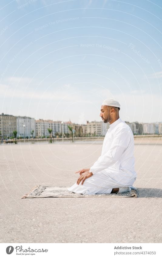 Muslim man praying on beach muslim arab islam religion tradition culture rug ritual worship holy male ethnic authentic sand arid desert blue sky cap robe faith