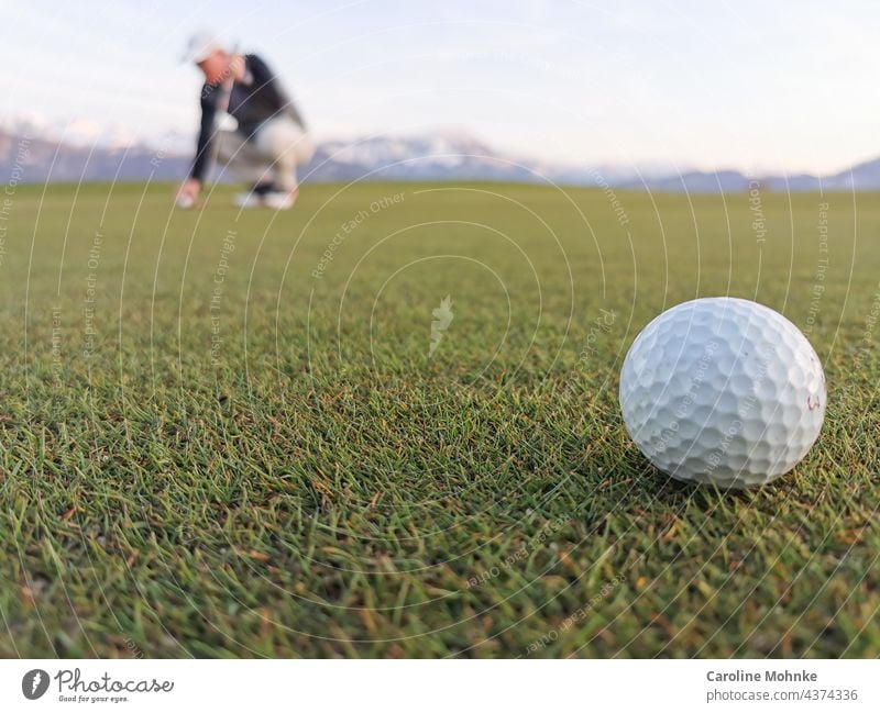 Golf ball on a green - in the background a golfer aligning his ball in front of an alpine panorama Golfer Nature Sports Golf course Leisure and hobbies Playing