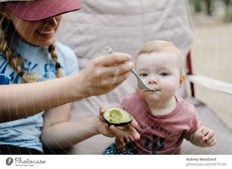 mom feeding her baby daughter an avocado outdoors while camping blonde blue eyes california camper campsite caucasian chair child childhood cooking eating