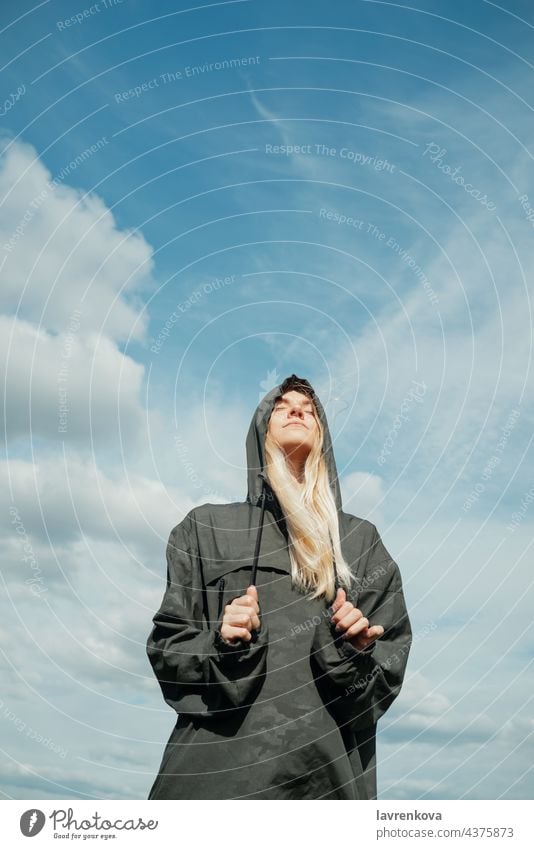 Young blond woman in khaki shirt with a hood standing in front of blue sky with her eyes closed pretty vertical landscape summer explorer freedom wind blonde