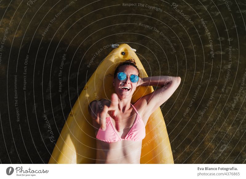 top view of happy caucasian woman lying on yellow canoe in lake during sunny day pointing with finger to camera.. summer time. Sports, adventure and nature. Woman in swim wear and sunglasses