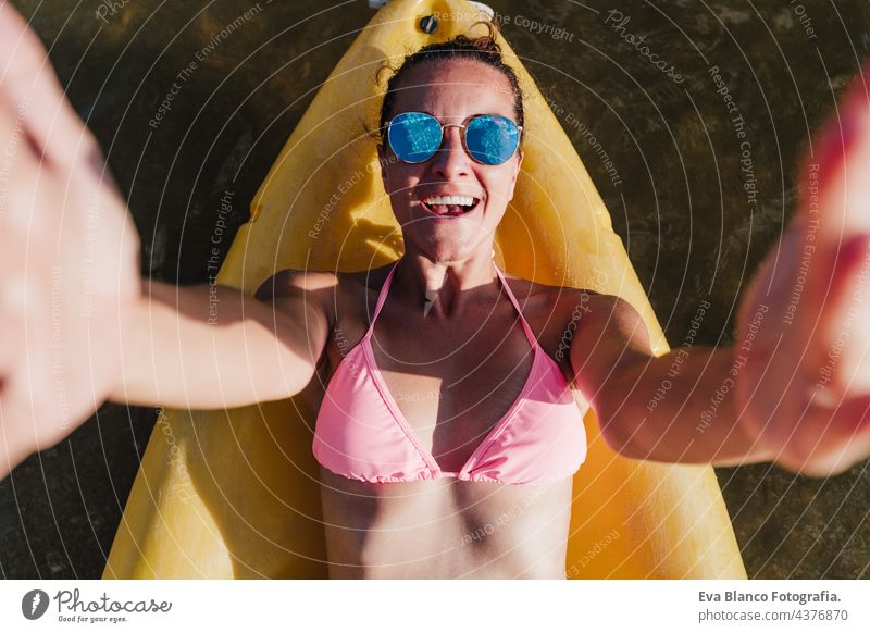 top view of happy caucasian woman lying on yellow canoe in lake during sunny day holding camera to take selfie.. summer time. Sports, adventure and nature. Woman in swim wear and sunglasses