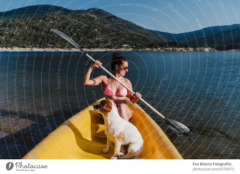 young caucasian woman and jack russell dog sitting on yellow canoe in lake during sunny day. Woman rowing. summer time. Pets, adventure and nature swim wear