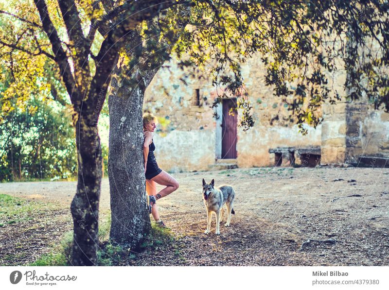 Mature caucasian woman standing outdoor with a wolf dog in a rural area with trees and a house. lifestyles people mature young women portrait adult ethnicity