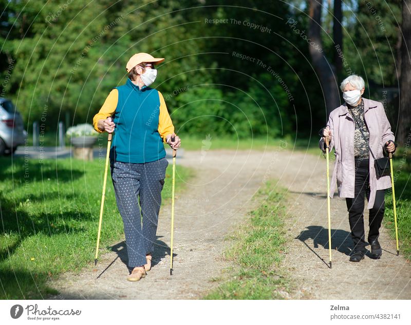 Two senior women wearing medical masks walking with nordic walking poles during covid-19 pandemic lifestyle leisure elderly women people training park fitness