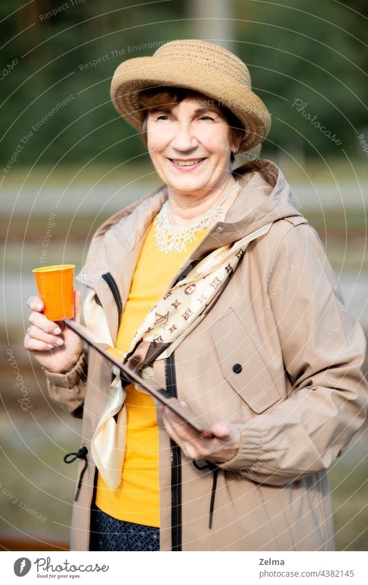 Smiling senior elderly woman with a hat, tablet and a glass of coffee traveling on during covid-19 pandemic old people face portrait smiling summer holiday