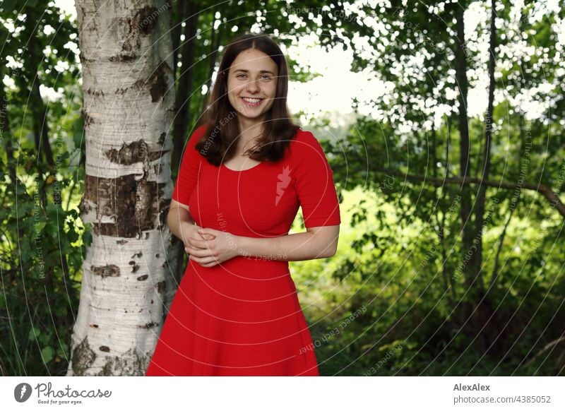 Young woman in red summer dress stands in the nature in the green and looks smiling into the camera Woman see Green Nature Brunette Dress red dress Grass