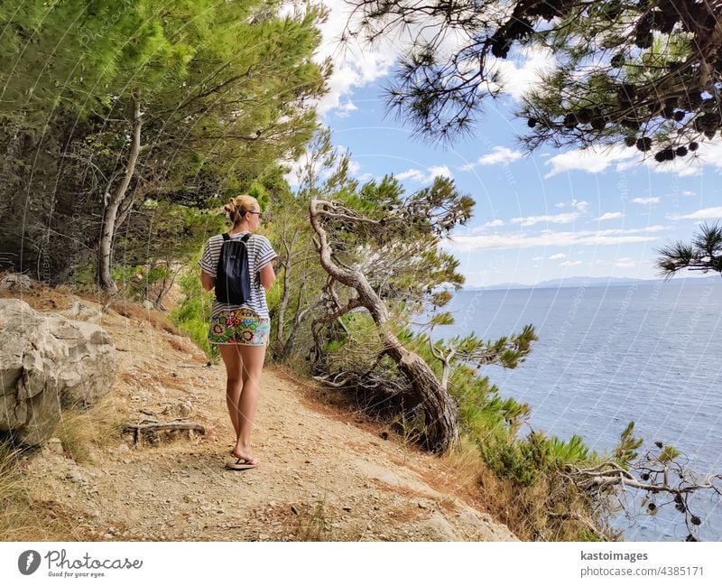 Young active feamle tourist wearing small backpack walking on coastal path among pine trees looking for remote cove to swim alone in peace on seaside in Croatia. Travel and adventure concept
