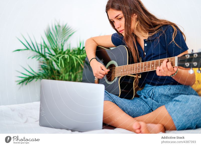 Young woman with laptop playing guitar music learn online at home acoustic practice female student young hobby lesson tutorial instrument watch education using