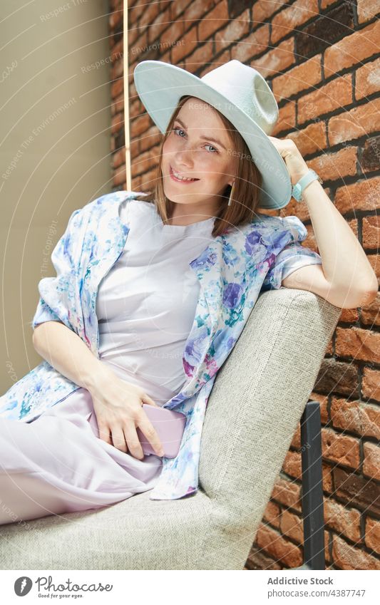 Trendy woman in hat resting on chair against brick wall smile style sincere friendly kind portrait smartphone gadget device sit cellphone rough stylish ornament