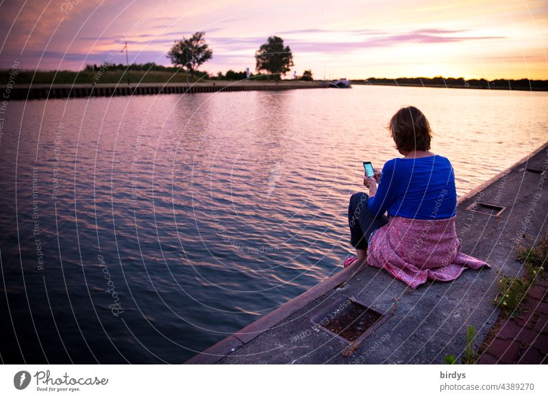 Woman sitting on a quay wall at sunset looking at her smartphone Water Sunset Landscape Horizon Sunlight free time Sit sneeze Twilight River evening sky
