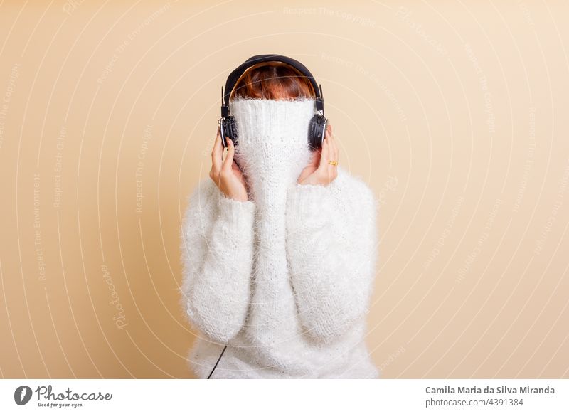 Fun portrait of girl with her trendy white sweater over her head hiding, cold. Listening to music with headphones. Woman with tied hair. Photo in studio. adult