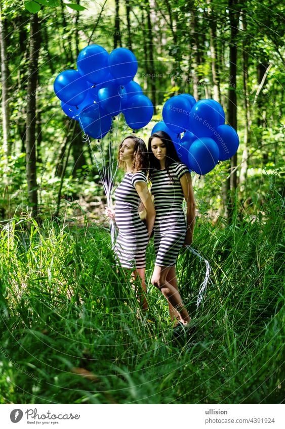 two women in black and white striped dress stand behind each other with blue balloons in the forest. artistic photography, copy space bunch helium free hold
