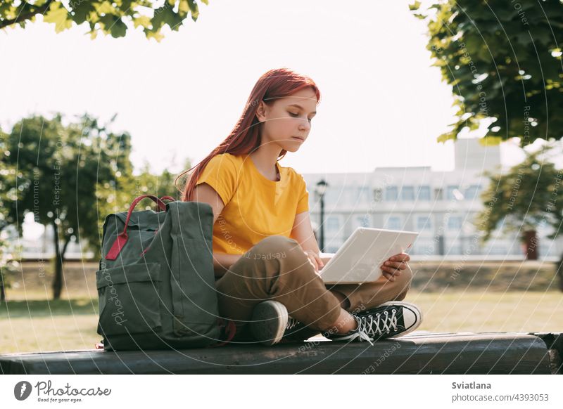 A teenage girl is sitting on a park bench with a laptop and preparing for lessons or exams. The concept of training and education backpack using student woman