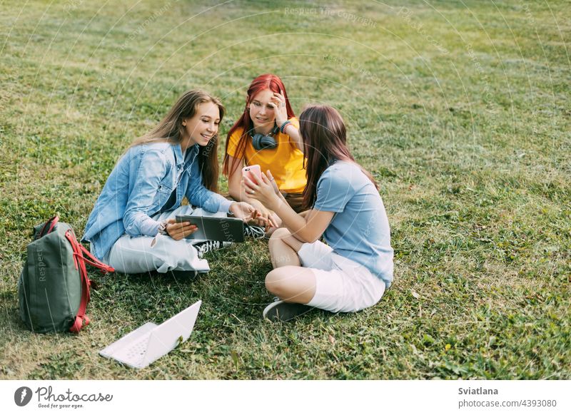 Teenage girls are sitting on a green lawn in a park with a backpack and a digital tablet and are preparing a joint project. Education and knowledge, lifestyle of teenagers