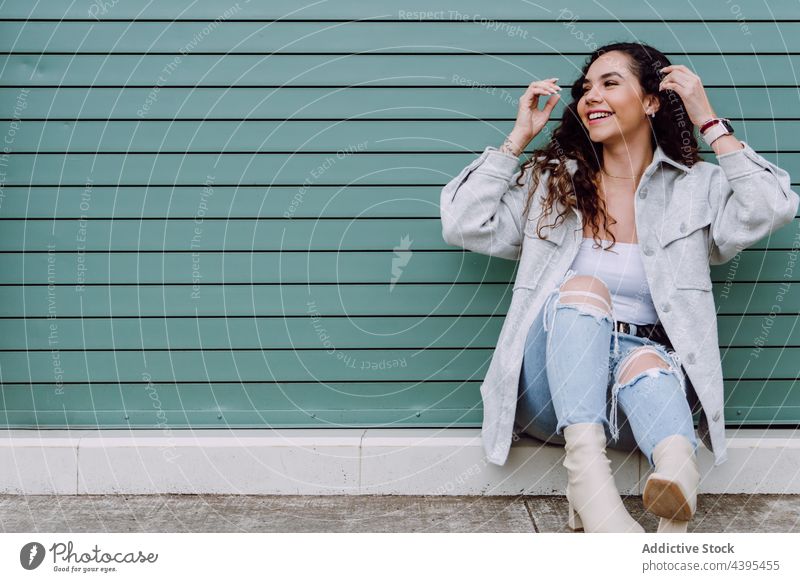Glad woman sitting near building wall in city toothy smile charming style street urban trendy fashion appearance female lean on hand cheerful glad outfit happy