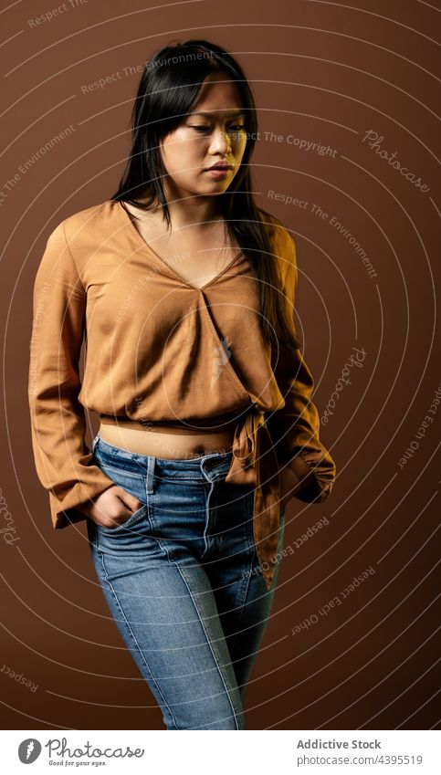 Serious Asian woman in stylish outfit in studio looking down style apparel model delight trendy appearance female asian ethnic smile positive jeans young