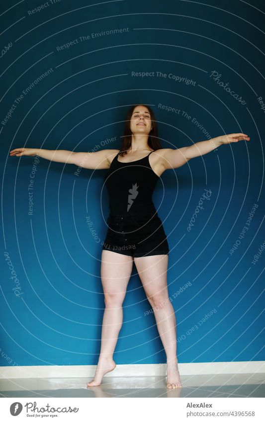 Young woman in black tank top and hot pants stands barefoot in front of a petrol blue wall and raises her arms in a dance pose Brunette Athletic 18 - 30 years