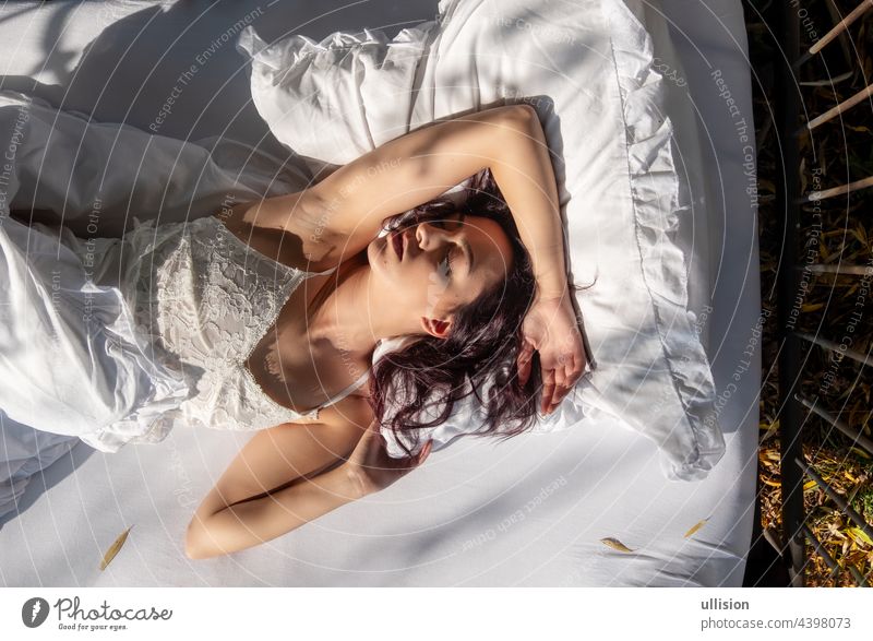 Portrait of an attractive, contented, young, sexy dark brown haired woman lying relaxed in bed, enjoying and snuggling with fresh, soft, white sheets and pillows in the morning sun, copy space