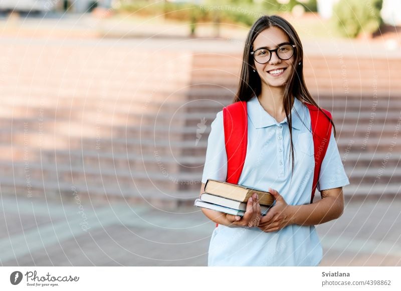 A smiling student girl with glasses, books and a red backpack. Portrait of a student on a city background. The concept of education college stair street