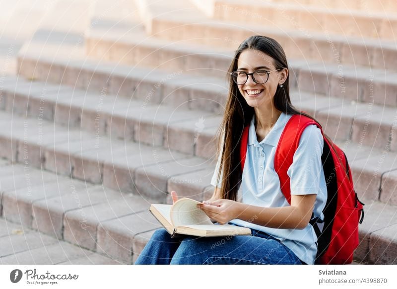 A pretty girl student in glasses, with a backpack is sitting on the stairs near the university, reading a book, preparing for lectures or exams. Training and education