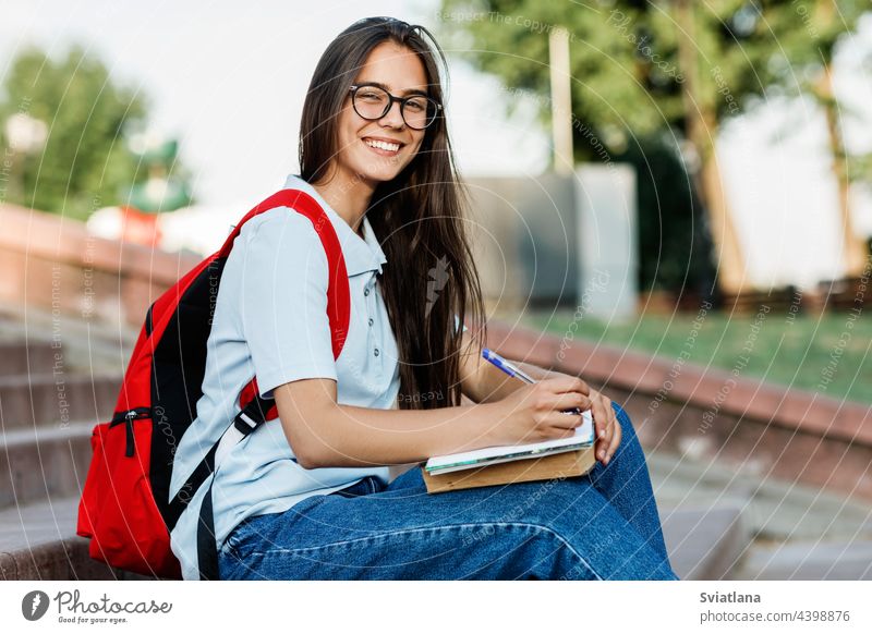 Portrait of a charming student girl with glasses sitting on the stairs in the city writing book notebook backpack focused notepad campus female education woman
