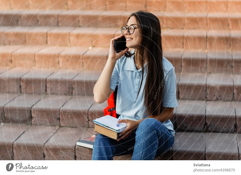 A cheerful girl is talking on a mobile phone with her friend, sitting with a notebook on the stairs in the campus learning smiling backpack outdoors notepad