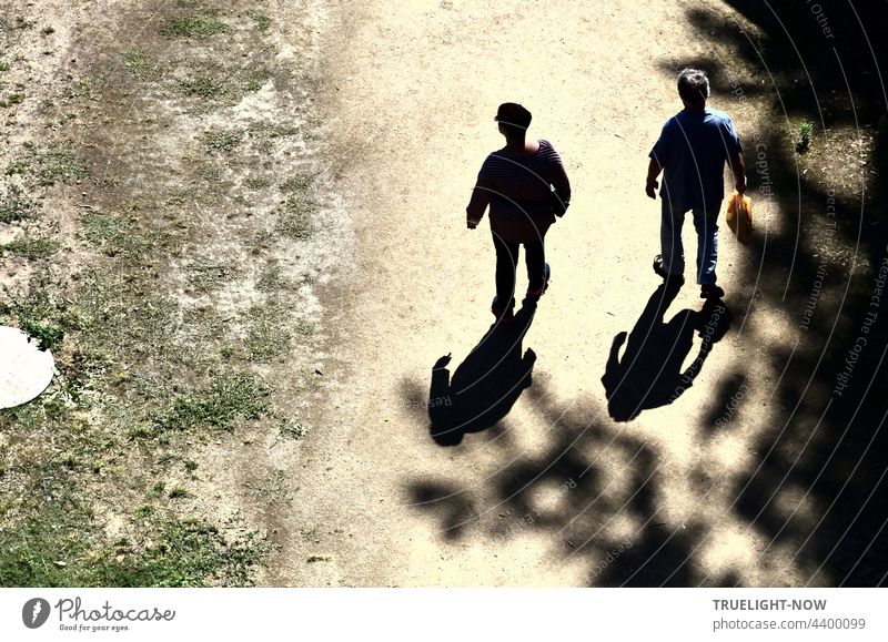 Two men walk side by side on a sandy path. Their shadows follow them relentlessly through the heat. One wears a cap on his head, the other a plastic bag in his hand. Tree shadows from the right and a pale meadow with a manhole on the left