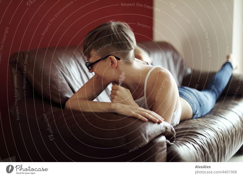 Young, slim woman with glasses in jeans and tank top lies turned away on the leather couch and laughs young maiden strappy top Jeans Couch along Decompose Skin