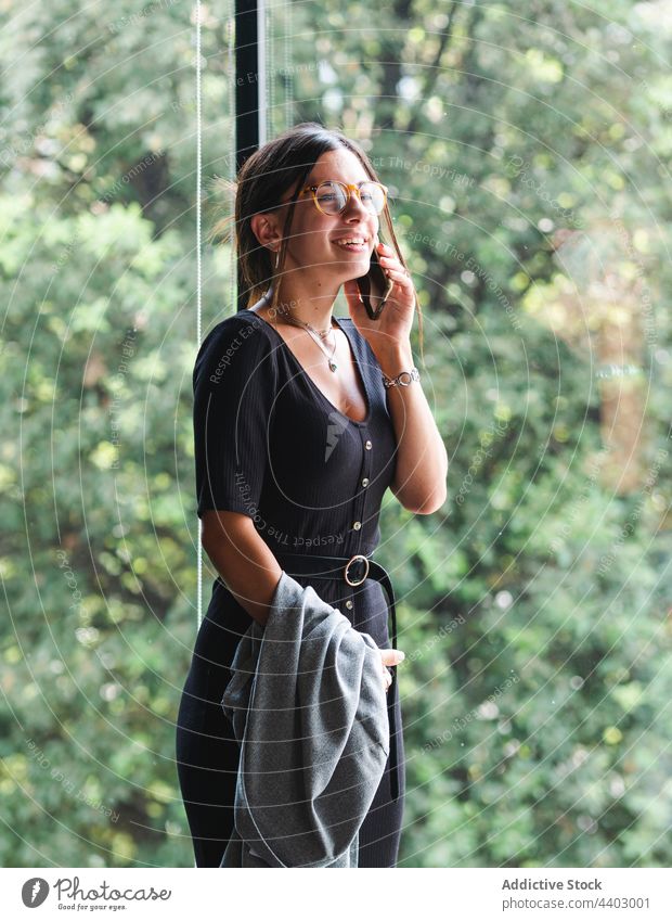 Cheerful woman talking on smartphone near window conversation mobile speak glass wall cheerful female smile style device gadget call connection building happy