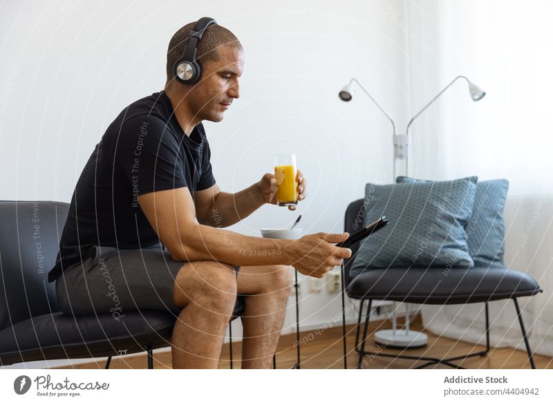 Content man drinking juice and using tablet at home watch video orange morning male browsing surfing sit connection gadget device internet online headphones