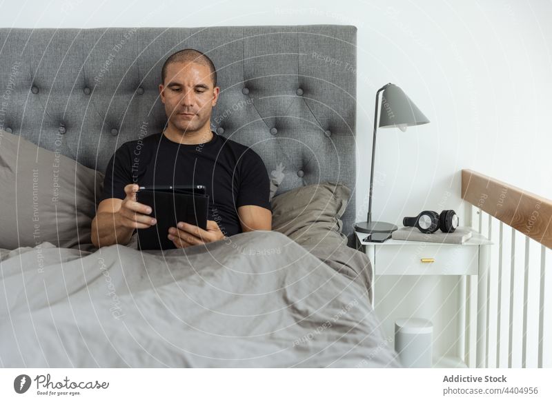 Man browsing tablet in bed in morning man awake bedroom using surfing online male home gadget device comfort internet addict early blanket chill serene sit calm