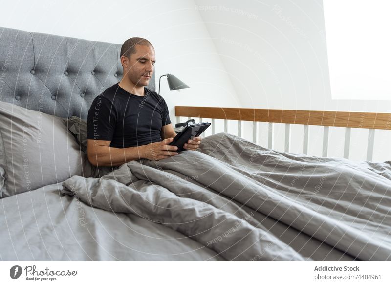 Man browsing tablet in bed in morning man awake bedroom using surfing online male home gadget device comfort internet addict early blanket chill serene sit calm
