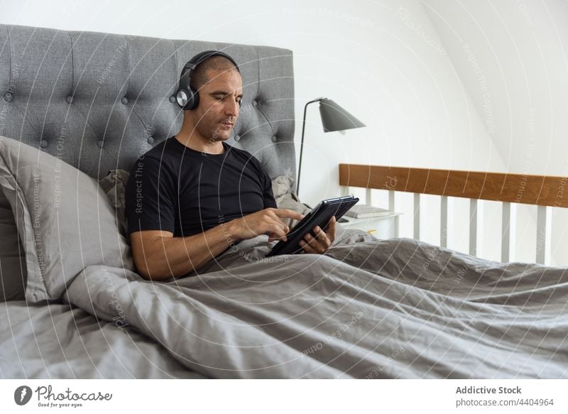 Man using tablet in bed in morning at home man browsing listen music bedroom awake male gadget rest device relax watch video surfing internet headphones