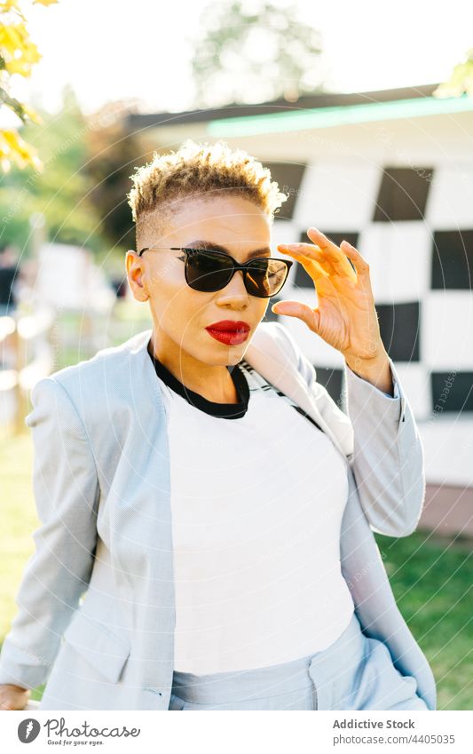 Black woman in stylish wear in sunshine sunglasses portrait individuality fashion style haircut park red lips black ethnic african american makeup decorative