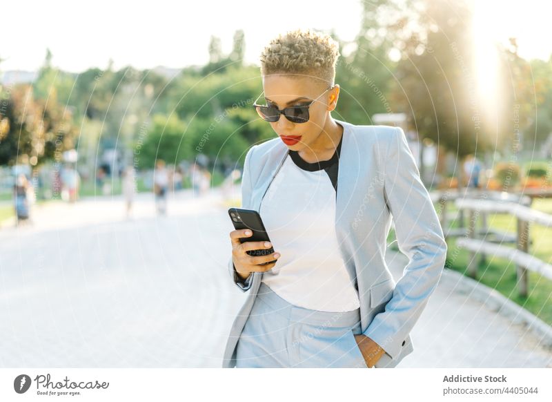 Trendy black woman chatting on smartphone while walking on pavement fashion style individuality sunglasses using gadget device cellphone text messaging watching